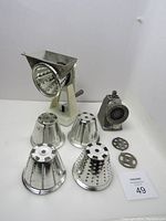 Photo showing the vintage manual Universal Dazey Mates meat grinder with 3 blades and salad maker with 5 heads alongside their metal base with suction, all displayed on a white surface.