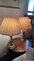 Pair of brass table lamps on a wooden round side table near a window, lit to show glow of pleated fabric shades.
