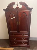 Front view of solid brown wood armoire with two paneled doors and three drawers below, brass tone handles, and curved top.
