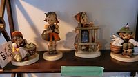Four ceramic Hummel figurines on a wooden shelf, showing full set.