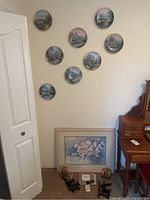 Eight decorative plates mounted on a wall showing scenic cottage and nature scenes, with a framed floral artwork and brass ship book holder on the floor below them.