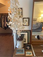 Clay whiskey jug with dried white branches and various framed pictures behind and beside it.