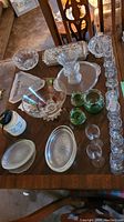 Full assortment of glass serving dishes, vases, and bowls displayed on wooden table, including various shapes and sizes, with floral etched designs and colored glass vases.