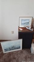 View of two framed nautical ship prints, one resting against wall, one standing on black pedestal; partial ship model visible on right.