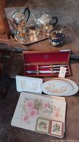 Photo of silver plate tea set items with ornate tray and coasters, Gerber carving set in wooden box, and floral placemats and coasters laid out.