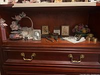 Display of vintage vanity set including hairbrushes, mirror, framed pictures, decorative flower basket, and small metal items on wooden cabinet.