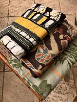 Stack of three folded tablecloths showing different patterns: batik style in black and yellow, printed multi-color cloth, and a green leafy design.
