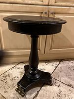 Full view of the black painted wood side table showing shape, pedestal, base, and brass ball feet.