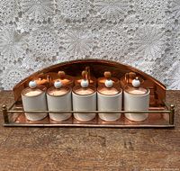 Front view of the copper spice rack with five ceramic and copper spice jars on wooden surface with lace background.