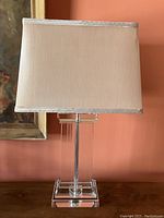 Cut glass lamp with double pedestal base and beige fabric shade sitting on wooden surface against orange wall.