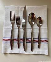 Flatware place setting shown on white cloth with red and blue pattern. Includes large and small forks, knife, large and small spoons in polished stainless steel with Splendide pattern.
