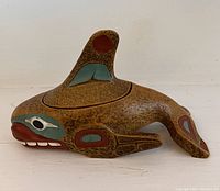 Side view of handcrafted whale box with painted facial and body markings in ochre red, green, white, and black colors on speckled wood texture.