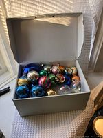 Box containing over two dozen vintage glass Christmas ornaments of various colors and reflective surfaces.