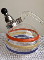 Front angle view of the round glass tea kettle with colorful banded stripes of orange, yellow, and blue, showing ribbed clear glass handle and aluminum top with black bakelite lid knob.
