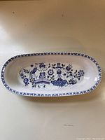 Top view of oblong serving dish showing detailed blue and white folkloric design with two figures and floral motifs, glossy finish, and decorative rim pattern.