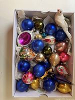 Box showing multiple glass ball ornaments in blue, green, gold, and pink colors, a white bird figurine at the top right, and various shaped vintage ornaments.