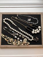 Top view of 5 vintage costume necklaces arranged on a black background, showing variety of beads and textures including faux pearls, glass beads, black and white Talbots beads, and Mother of Pearl style beads. Also shows 3 pairs of leaf-shaped earrings.