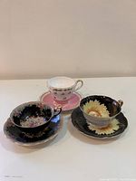 Three vintage teacups and saucers: black with yellow sunflower, black with petite floral, and pink with roses.