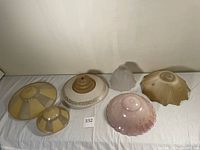 Six vintage lamp or light shades displayed on white cloth, showing various glass and plastic shades including pink, clear, and cream colors with different shapes and textures.