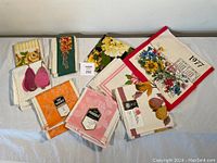 Image showing 12 linen tea towels with assorted floral, fruit designs and visible tags including Pedigree and hand printed marks