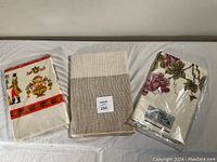 Three folded tablecloths on a white surface, including one with a colorful print, one beige and cream weave, and one labeled Grape Arbor in packaging.