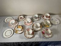 Full set of ten bone china teacups and saucers with various floral and gold designs arranged on a white cloth surface.