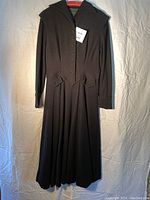 Full front view of vintage black dress hanging, showing collar, buttoned front, waist tabs, and long sleeves.