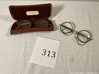 Two pairs of vintage eyeglasses; one pair in a hard brown case and one pair with round black rims and silver temples lying next to it.
