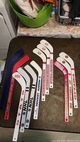 Twelve mini hockey sticks laid out showing different brands and teams including NHL, State, and Hockey Nation.