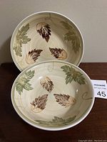 Two ceramic serving bowls stacked, showing internal leaf pattern decoration in brown and green on cream.