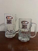 Two A&W glass mugs side by side against a neutral background, showing front logo and handle detail.