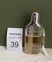 Glass bottle of Burberry The Beat Eau De Parfum with silver top and attached strap displayed against dark background next to MaxSold lot number 39