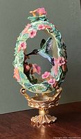 Front view of the Franklin Mint Faberge hummingbird egg on gold-colored stand with floral and hummingbird details.