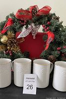 Four white ceramic mugs with embossed holiday garland pattern and gold rims, displayed in a row with a holiday wreath and red bow in the background.