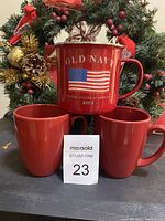 Red metal Old Navy 2001 mug with American flag design and two red stoneware mugs behind it on a table in front of Christmas wreath decorations.