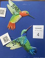 Two metal hummingbird hanging decorations displayed on a blue background with their tags visible. One is green and orange colored, the other is green and blue.