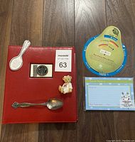 Overview of the full lot including all items on a wood floor: red recipe binder, two silver spoons, Cherished Teddies magnet, recipe cards, and cheese/wine pairing card.