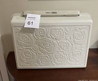 Full front view of the cream hard carrying case with embossed rose design and handle on top.