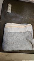 Folded grey fleece blanket with beige and white striped edge visible on a dark surface.