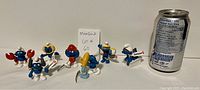 Eight Smurf figurines arranged with soda can for scale. Characters include one with crab claws, one pouring from jug, and several in sporty or Zodiac themes. Tags attached.
