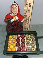 Front view of the female caroler figurine with open mouth and holding lemons next to a ruler for size reference, showing the cart full of miniature fruit.