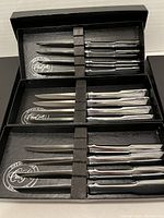 Three black boxes each containing four stainless steel steak knives lined up neatly inside with logo visible in the box lining.