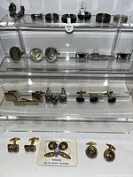 Several tie clips and cufflinks displayed on clear acrylic tiered stands showing variety of designs and metals.