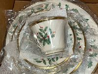 Five piece place setting wrapped in plastic in original box showing all plates and cup with holly and berry holiday design and gold trim.
