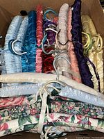 Box of satin and cotton covered padded hangers in various colors including light blue, red, purple, beige, gold and floral print. Several hangers have metal skirt/trouser clips attached.