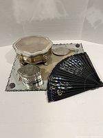 Photo shows the mirrored picture frame lying flat with the black folding fan partially open, silver-tone octagonal jewelry casket, silver-tone oval Madonna rosary box, and small handheld oval souvenir mirror placed on the frame.