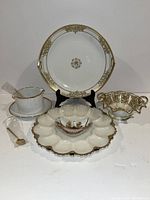 Front view of various vintage hostess tableware including milk glass deviled egg tray, gold patterned plates, cups, and small serving pieces arranged on white background