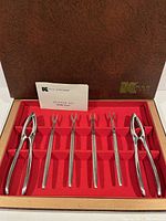 Full view of shellfish tool kit including 2 lobster crackers and 6 seafood forks in red velvet-lined box with brown exterior and instruction card.