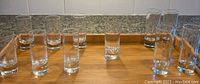 Photo of 11 transparent glass shot glasses arranged on a wooden tray, 7 shorter and 4 taller ones, all clear glass in good condition.