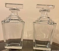Photo of two clear crystal whisky/liquor decanters with square stoppers and geometric design on reflective surface.